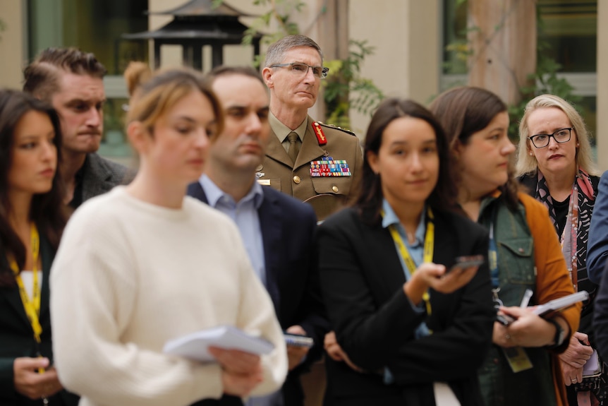 Campbell in military uniform looks out of frame, a row of journalists in front of him.
