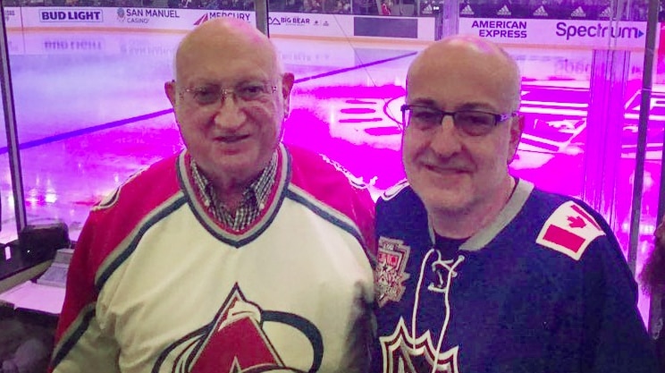 Two men stand arm in arm, and smile at the camera with an ice hockey field behind them.
