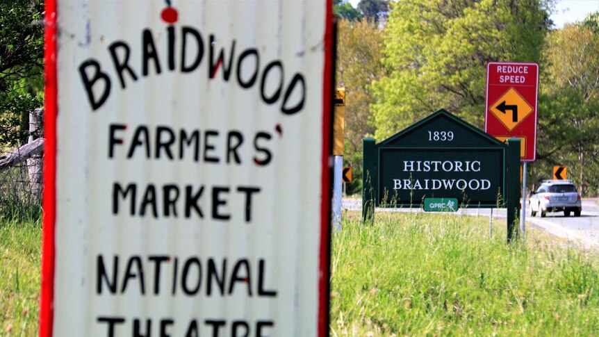 Close up of sign advertising events on in Braidwood, with 'Historic Braidwood' town sign in background, next to road.