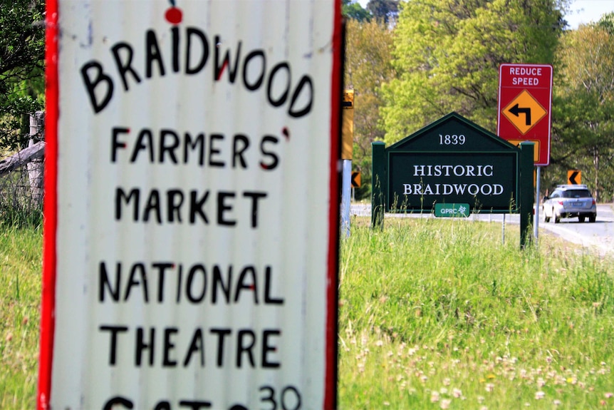 Close up of sign advertising events on in Braidwood, with 'Historic Braidwood' town sign in background, next to road.