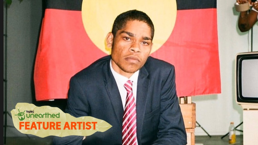 Rapper JK-47 kneels in front of an Aboriginal flag wearing a suit.