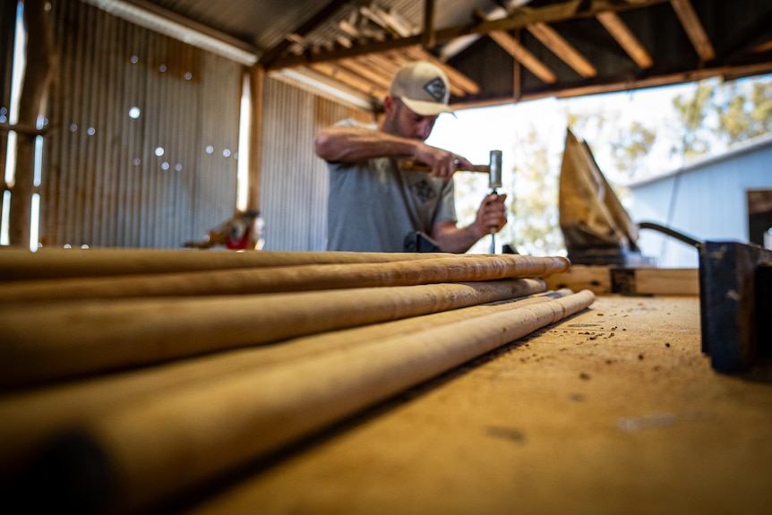 A man works with his tools in the background of a long working station with piles of wood cylinders on top. 