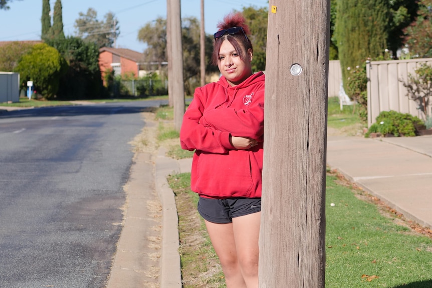 A young woman wearing a red hoodie and black shorts stands next to a telegraph pole on a suburban street.