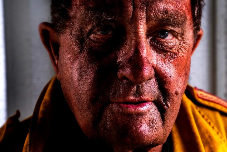 A close up of an older man's weathered face. He's in a yellow firefighting uniform. His uniform and his face is blackened by ash