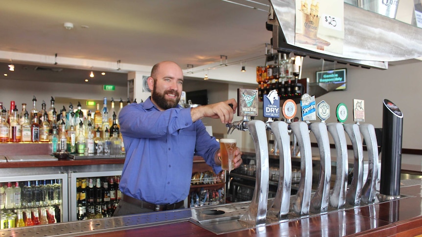 A bar manager wearing a blue shirt stands behind a bar and pours beer into a glass from a tap.
