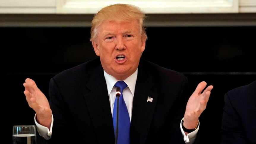 US President Donald speaks firmly with hands in the air wearing a black suit with a blue tie.