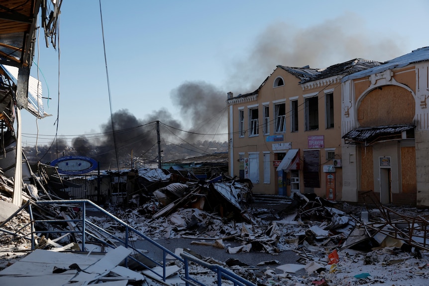 Smoke rises from a building while ruins of shelling on a clear day lie in the streets.