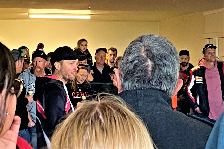 A man in a black cap and a black and red hoodie stands in a packed room speaking. It is lit by yellow light.