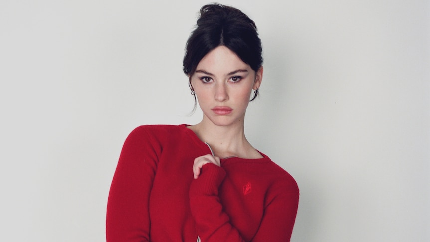 Promo photo of Gracie Abrams in a red jumper with her hair pulled up into a bun