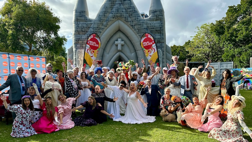 dozens of couples in wedding costumes posing outside of an inflatable church in a park