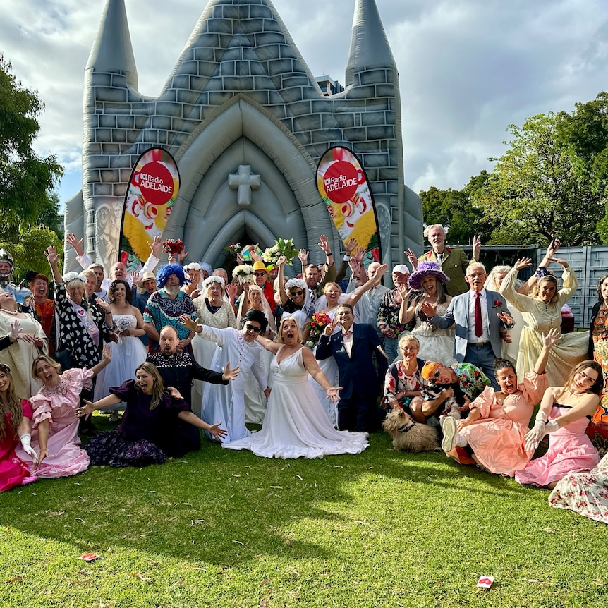 dozens of couples in wedding costumes posing outside of an inflatable church in a park
