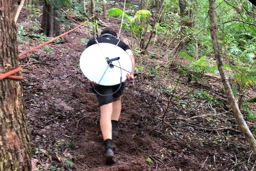 A man with a small satellite on his back walking up a steep mudding hill in the forest