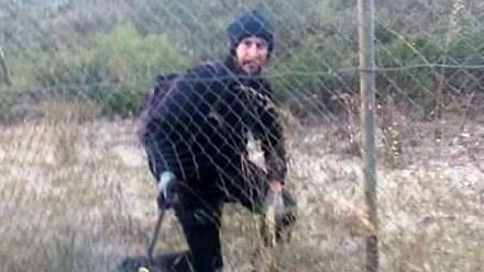 A man dressed in black climbing through a hole in a cyclone wire fence.