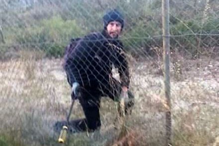 A man dressed in black climbing through a hole in a cyclone wire fence.