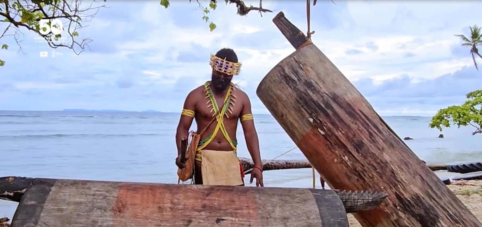 Man dressed in traditional attire beats drum. Ocean behind him. 
