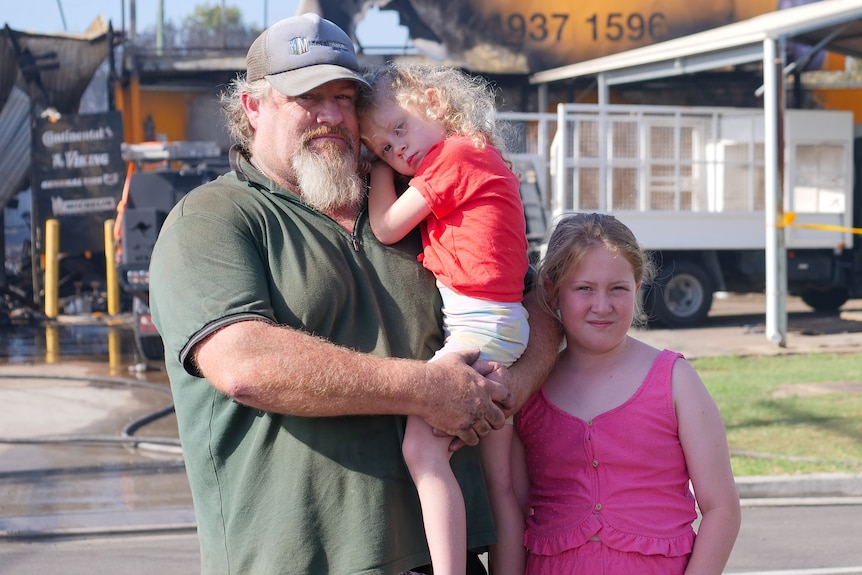Un hombre sosteniendo a una niña con otra niña parada a su lado, frente a una tienda de llantas quemadas.