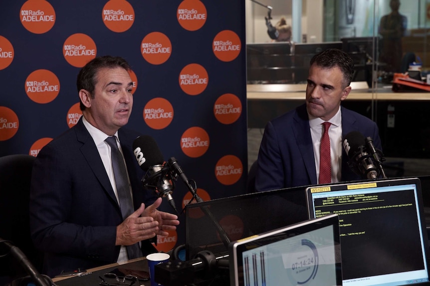 Two men in a radio booth with one one man speaking while the other looks at him.