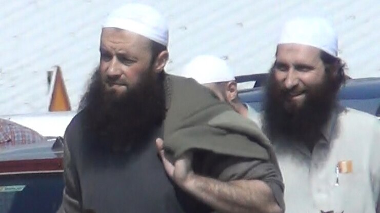 Three men with beards, two wearing white taqiyah caps, walk past a car