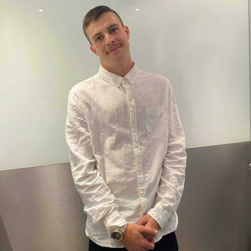 A young man stands in a white shirt smiling to the camera.