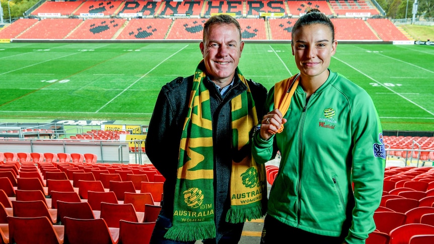 Alen Stajcic and Chloe Logarzo wear Matildas scarves as they stand on the terraces of Penrith Stadium with the field behind them