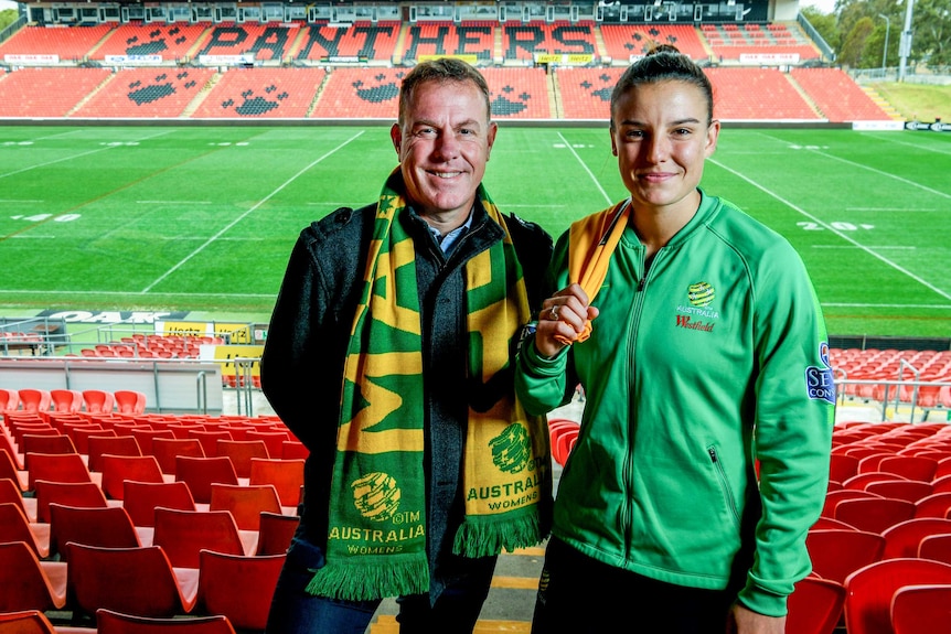 Alen Stajcic and Chloe Logarzo wear Matildas scarves as they stand on the terraces of Penrith Stadium with the field behind them