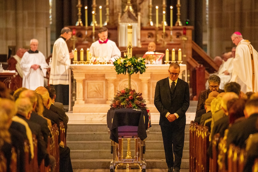 A funeral attendant stands with his hands clasped and head bowed beside the coffin while priests are on the altar
