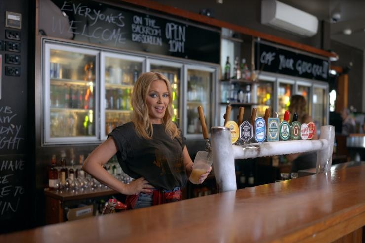 Kylie Minogue smiles as she pours a beer from a tap at a bar.