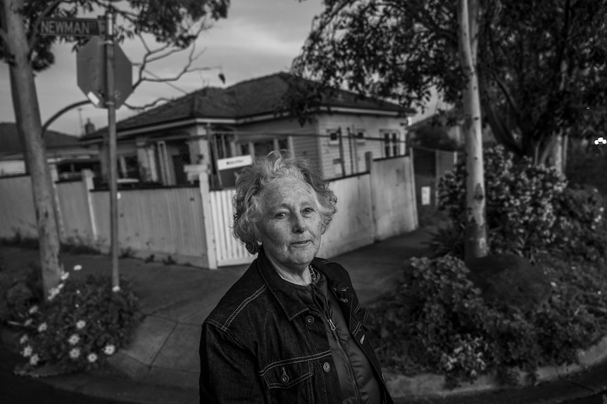 Elderly woman standing in front of a small house.