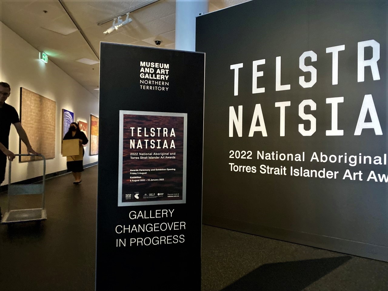 A sign saying 'gallery changeover in progress, with two people carrying boxes and painting on walls behind it.