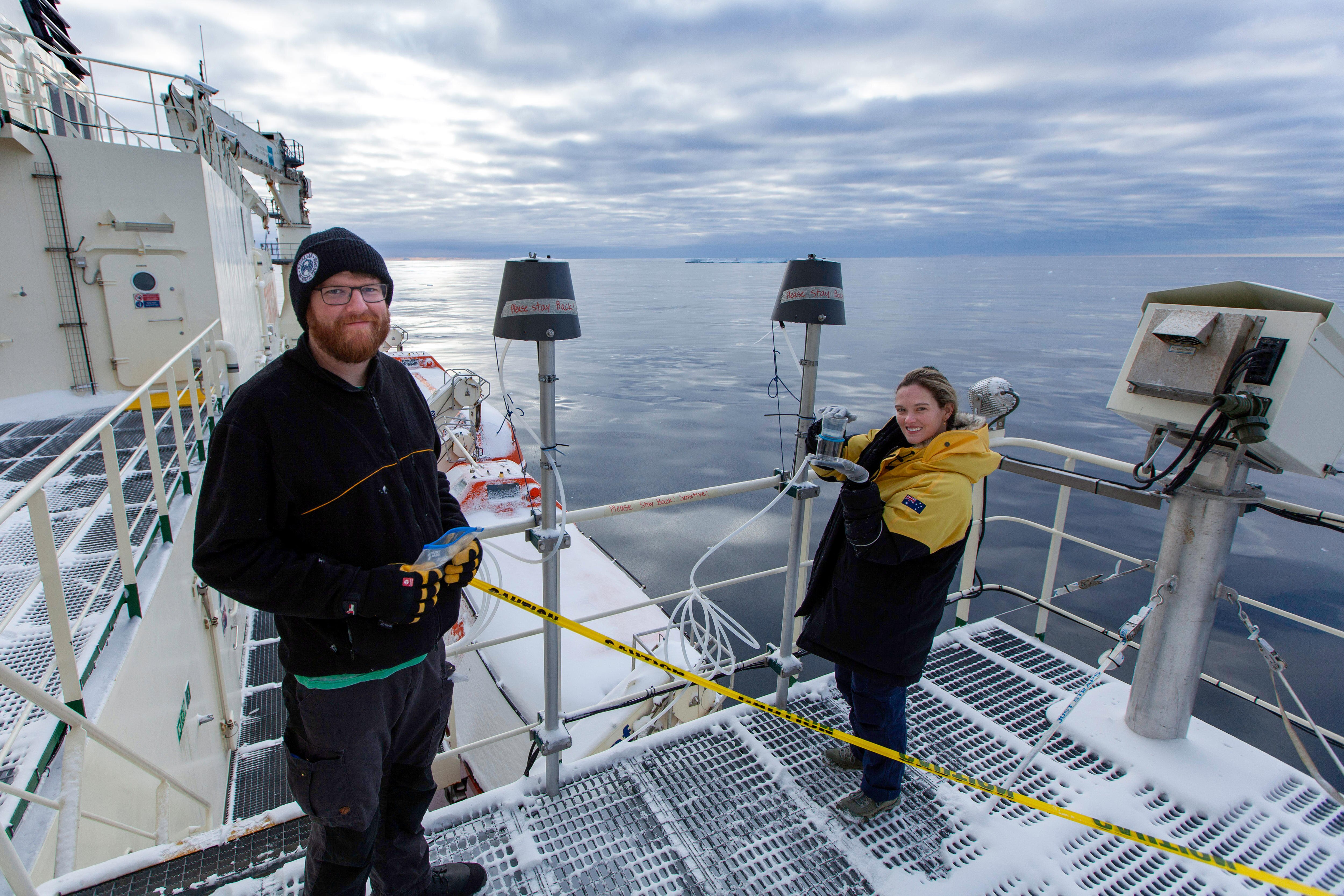 A man wearing black and a woman in a black and yellow coat stand on the deck of a ship. She is holding plastic containers.