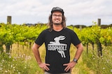 A man with long brown hair, a dusty cap, glasses and a black t-shirt stands smiling in the middle of a vineyard on a sunny day.
