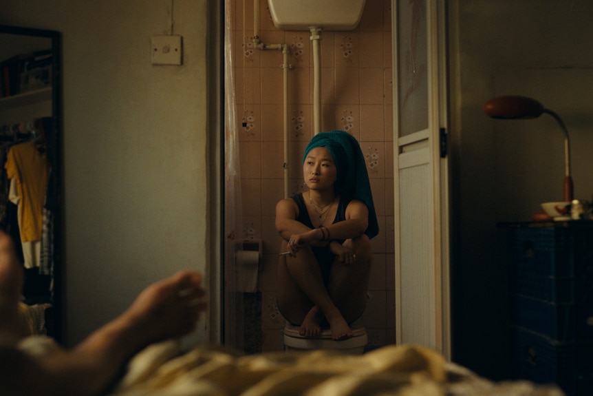 A TV still of Ji-young Yoo sitting on a closed toilet lid. She has her hair up in a towel.