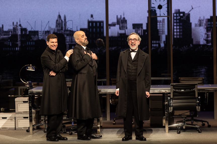 A scene from a stage featuring three male actors wearing black tie and long black coats