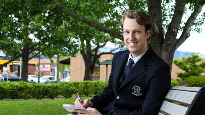 A year 12 high school student in uniform sits on a park bench taking notes