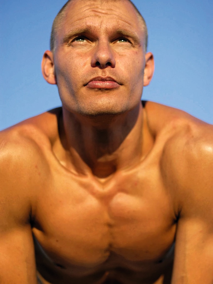 A shirtless toned and tanned middle-aged man with a shaved head and blue eyes looks into the distance. 
