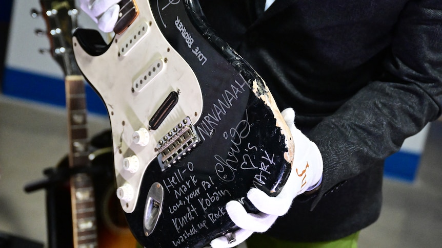 A man wearing white gloves is holding up the bottom of a Fender Stratocaster (guitar). It is signed in white.