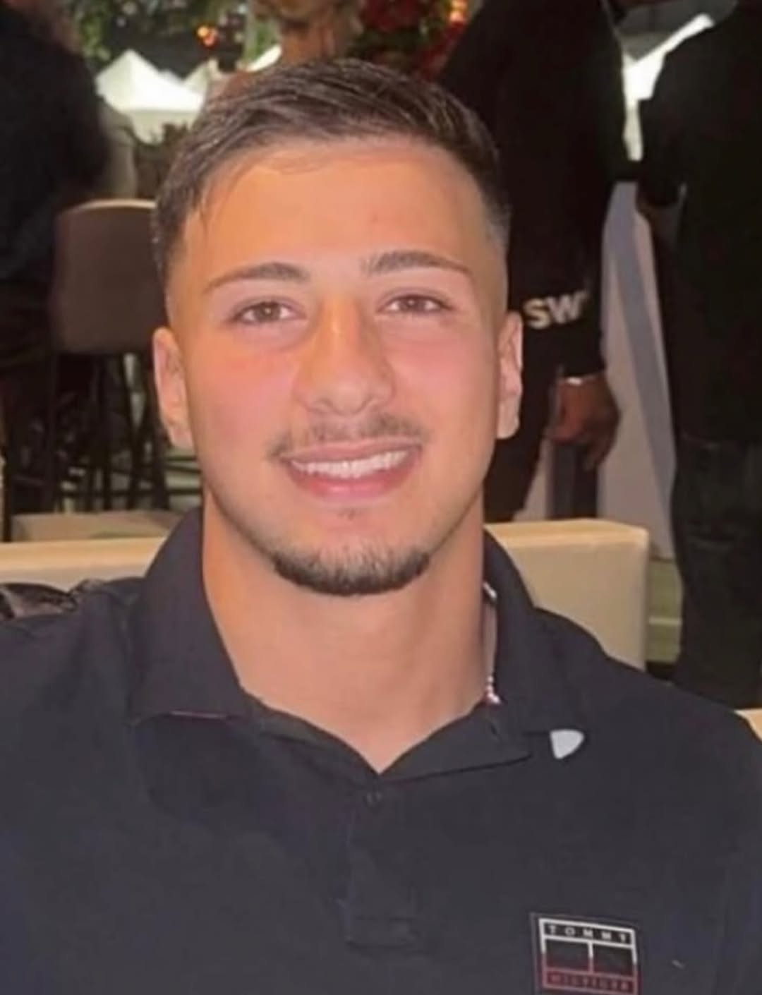 A young man with dark hair in a black polo shirt smiles looking directly at the camera.