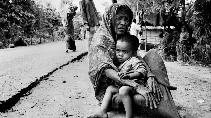 A Rohingya woman sits on the dusty ground with her grandchild in her lap.