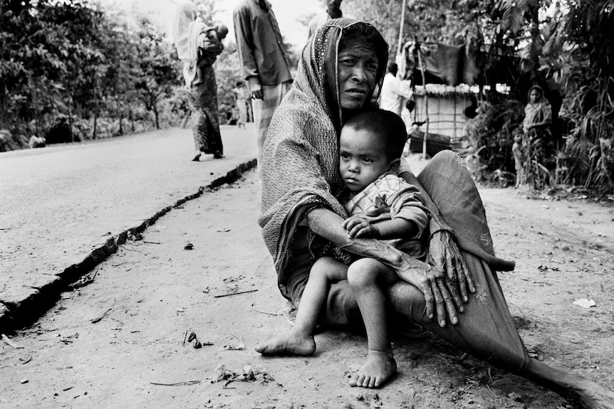 A Rohingya woman sits on the dusty ground with her grandchild in her lap.