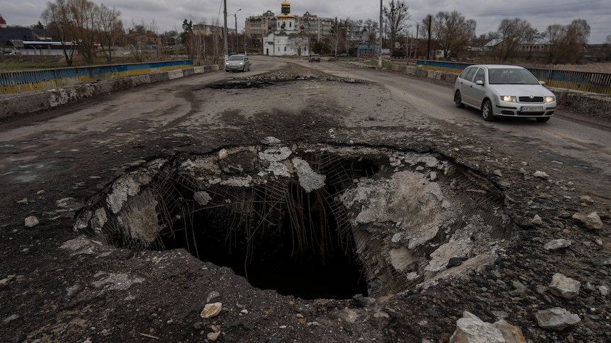 A large hole in the ground of a road with a car driving past.