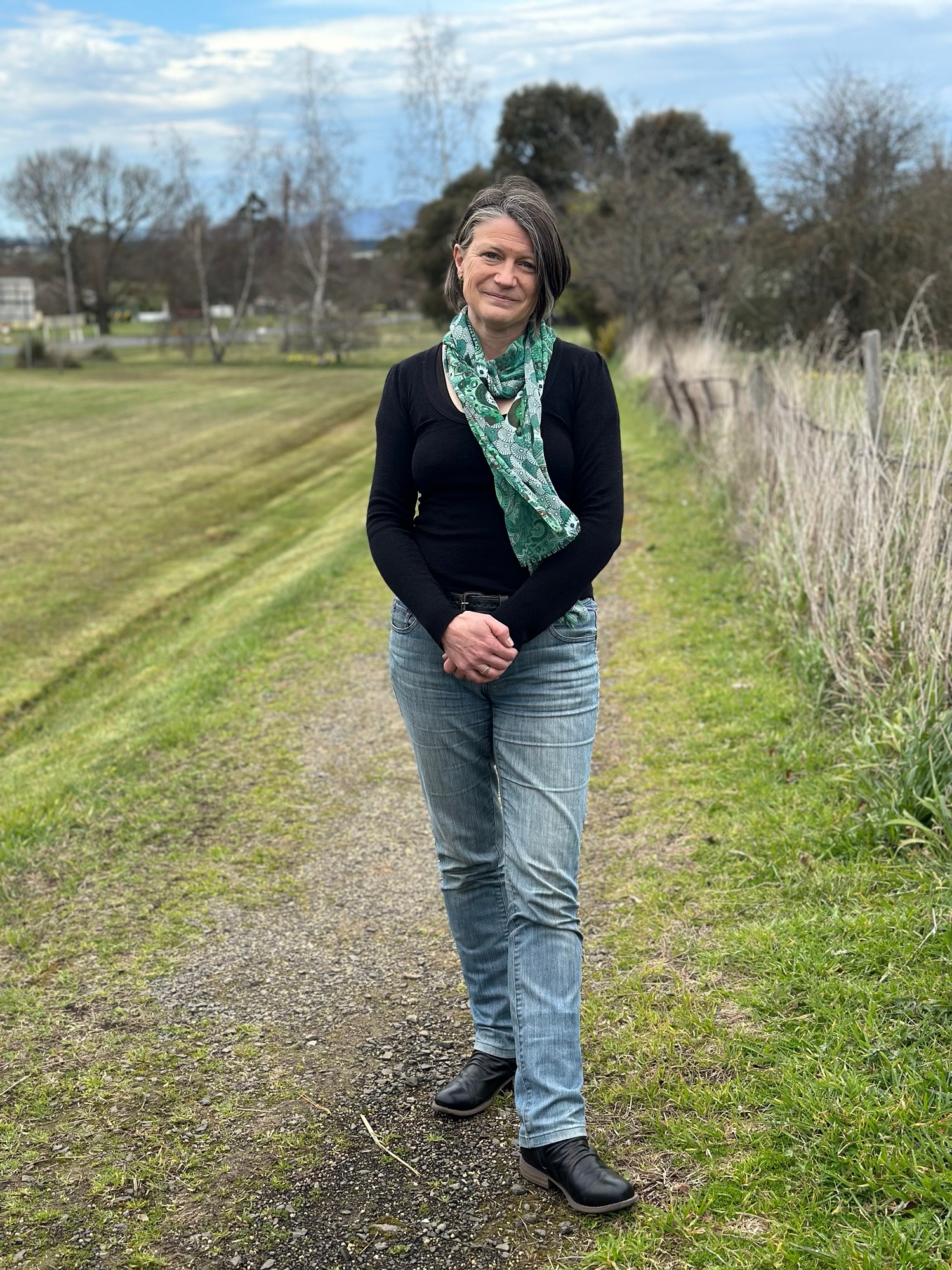 A woman with grey hair, black long sleeve shirt and a green scarf, stand in a paddock facing the camera. 