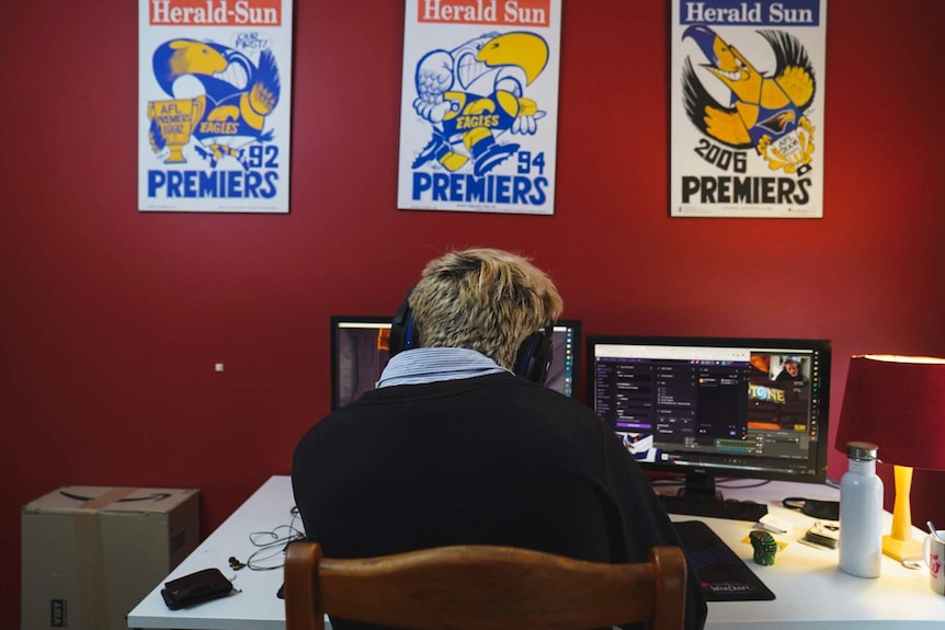 Alex Ridley sits at his computer with his back to the camera and three West Coast Eagles premiership posters on a red wall.