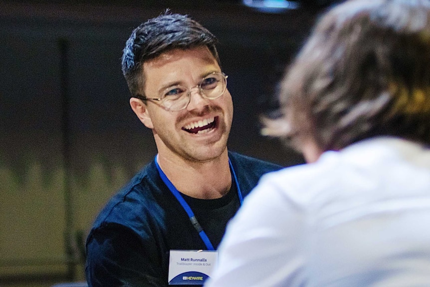 Matthew Searle sits left, smiling as he speaks with a person with their back to him and Matthew Runnalls, right.