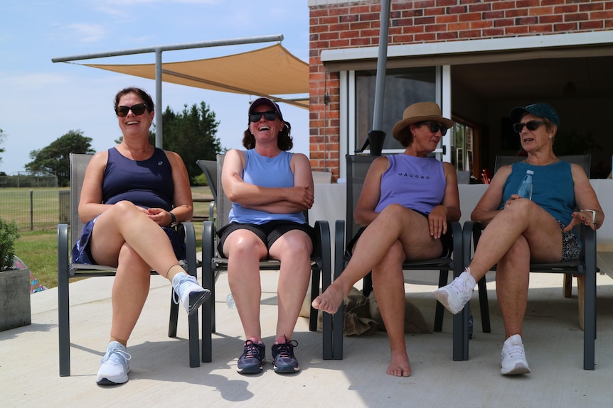 Four women sit on chairs on a deck in front of tennis clubrooms
