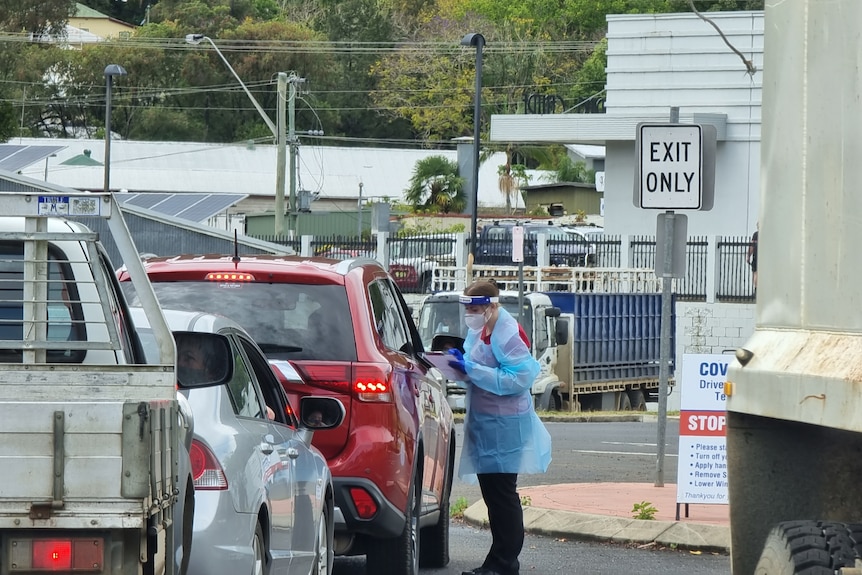 A woman wearing full personal protective equipment stands next to a car, with other cars queued behind it. 