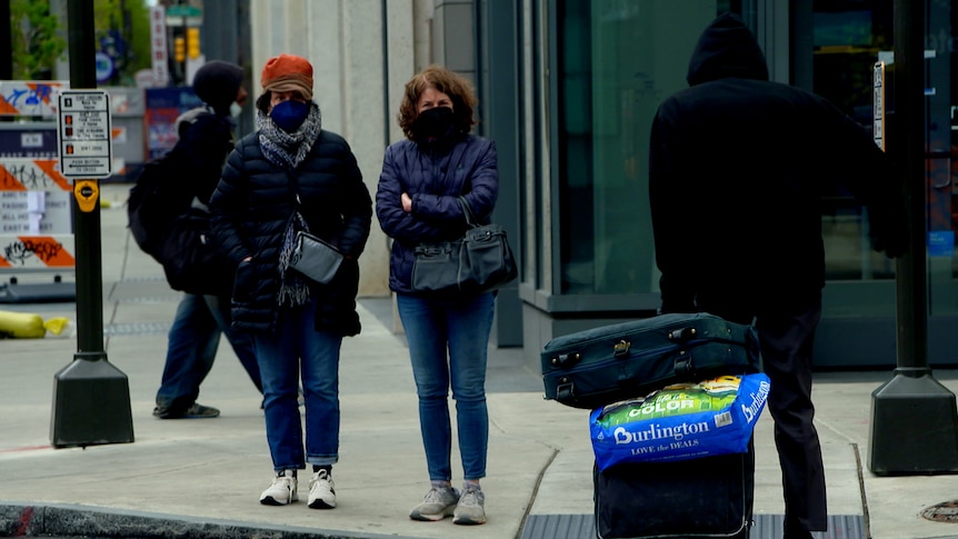 Two women in coats wait to cross the street while wearing masks