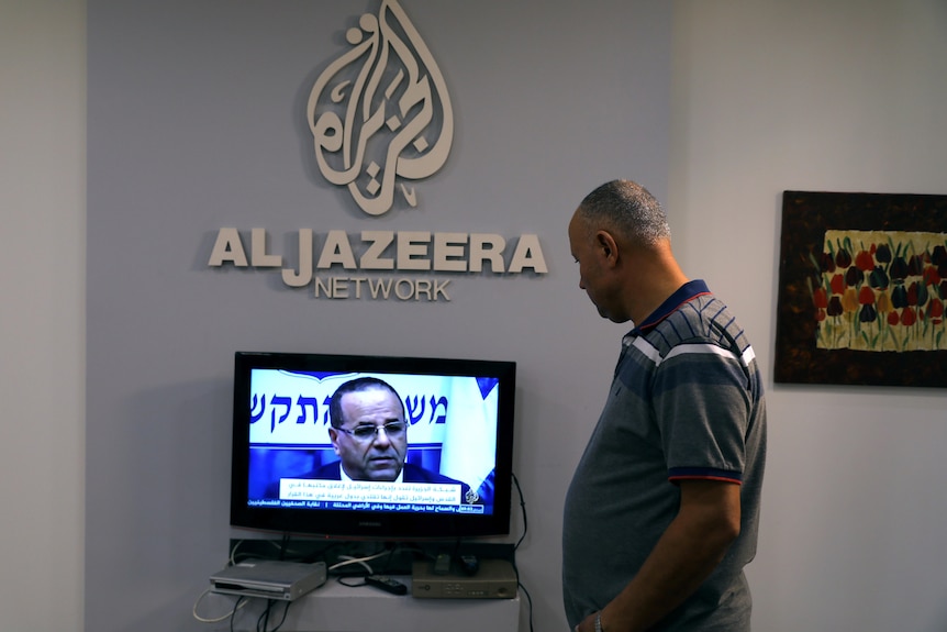 A middle-aged man in a grey polo shirt watches a small TV set up on a table underneath a large Al Jazeera logo on the wall.
