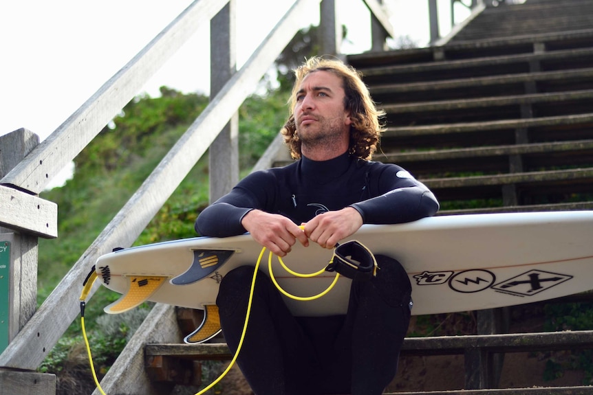 Indigenous surfer Jordie Campbell sits on steps in a wetsuit, holding his surfboard looking out to sea.