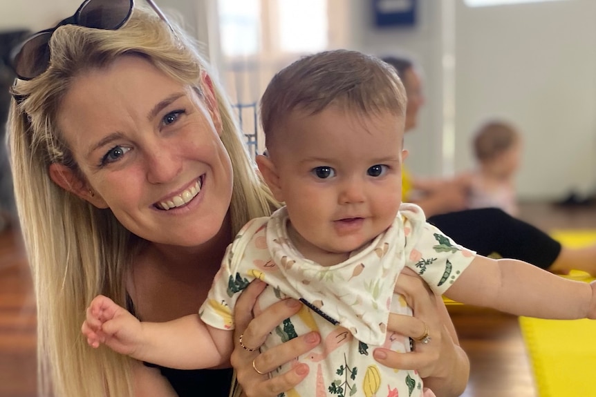 A young, smiling, blonde woman smiles holds a baby and looks at the camera, her sunglasses are perched on her head.