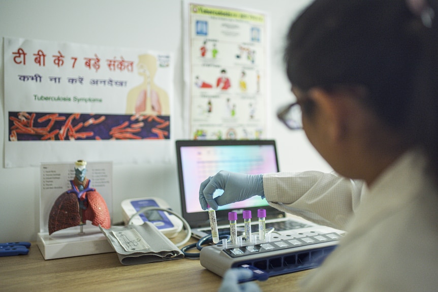 A woman in a lab coat puts test tubes in a device attached to a laptop.